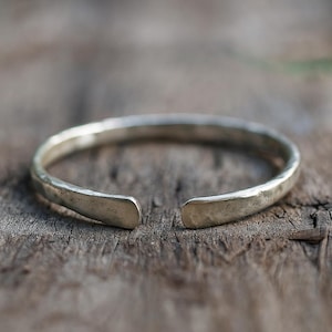 Op de afbeelding: Een zilverkleurige manchetarmband met een gehamerde textuur. Het open ontwerp maakt het gemakkelijk om te dragen. De armband wordt getoond op een houten oppervlak, wat de metaalglans benadrukt. Een eenvoudig, elegant sieraad.