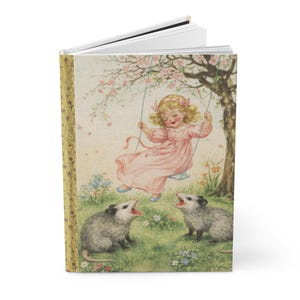 May include: A hardcover journal with a vintage illustration. The cover features a young girl in a pink dress on a swing under a flowering tree, with two opossums below. The spine is gold with a floral pattern.