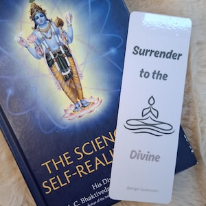 May include: A book titled "THE SCIENCE OF SELF-REALIZATION" with an illustration of a deity, next to a white bookmark. The bookmark reads "Surrender to the Divine" with a yoga pose graphic. The background is a fluffy, off-white surface.