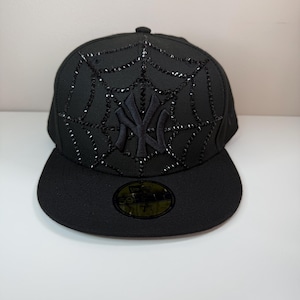 Puede incluir: Gorra de béisbol negra con diseño de telaraña y adornos negros. La gorra tiene una visera curva y el logo de los Yankees de Nueva York en negro. Es un estilo ajustado.