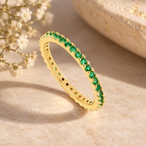 May include: A gold-toned band ring featuring a continuous row of small, round, vibrant green stones set along the outer edge. The inner surface of the band is clearly stamped with the text 925, indicating the metal composition of the jewellery piece.