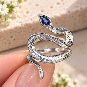 May include: A silver-coloured ring shaped like a coiled snake with a textured, scale-like pattern along its body. The snake's head is pointed upwards and features a single, small, pear-shaped dark blue gemstone set as the eye of the serpent.