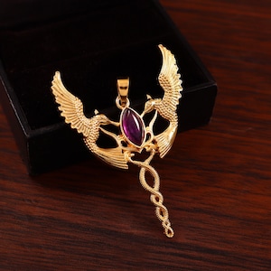 May include: Gold-coloured pendant featuring two stylised birds with outstretched wings flanking a central, oval-shaped purple gemstone. The pendant has an ornate, detailed design with a flowing, twisted tail.