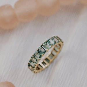 May include: An eternity band ring crafted from yellow gold, featuring a continuous row of emerald-cut, light green gemstones set in a prong setting. The ring displays a uniform arrangement of rectangular stones that encircle the entire band.