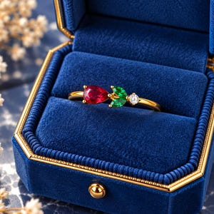 Pode incluir: Um anel de ouro com uma pedra vermelha em forma de pera, duas pedras verdes de corte marquise dispostas como folhas e uma pedra redonda transparente. O anel é exibido dentro de uma caixa de joias de veludo azul profundo com bordas douradas.
