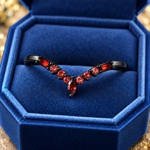 May include: A V-shaped ring crafted from black metal, featuring a central marquise-cut red gemstone, flanked by round-cut red stones and rectangular-cut red stones along the band, presented inside a deep blue velvet jewellery box.