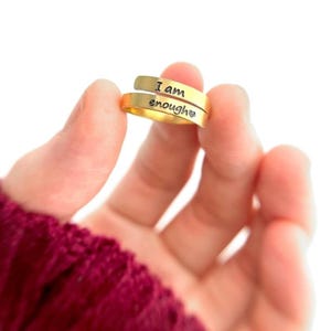 Peut inclure: Une bague dorée avec les mots "I am enough" gravés. La bague est tenue dans une main sur fond blanc. La bague a un design enveloppant.