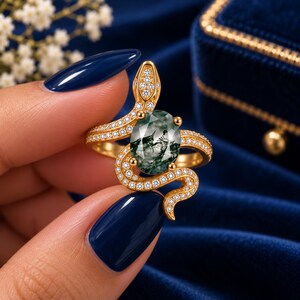 Puede incluir: Un anillo de oro con forma de serpiente enroscada con una banda y cabeza pavimentadas. El centro presenta una piedra preciosa de ágata musgosa de corte ovalado con inclusiones de color verde oscuro. El cuerpo y la cabeza de la serpiente están incrustados con pequeñas piedras blancas.