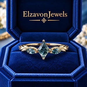 Op de afbeelding: Een gouden ring met een centrale vliegervormige blauwgroene edelsteen, geflankeerd door twee kleinere marquise-geslepen stenen op een band met bladvormige metalen details, gepresenteerd in een blauwe fluwelen doos met de tekst ElzavonJewels.