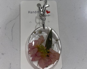 May include: Oval resin keychain with a pink flower and green leaf design. The clear casing is attached to a silver clasp. The tag reads "Handmade love".