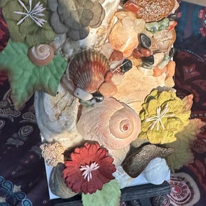 May include: A decorative rectangular art piece featuring a collage of seashells, pebbles, and artificial flowers. The artwork includes a variety of textures and colours, with a focus on natural elements. The piece is displayed on a small wooden easel.
