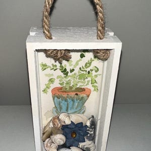 May include: A white wooden lantern with a rope handle. Inside, a watercolour painting of a potted plant and seashells. A blue flower and more shells are at the bottom.