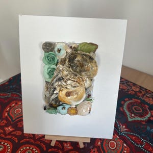 May include: A framed artwork with a collage of seashells, small stones, and artificial flowers in green, blue, and yellow. The artwork is mounted on a white background and displayed on a small wooden easel.