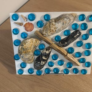 May include: A decorative piece featuring seashells, blue glass beads, and a wooden element arranged on a white background. The composition includes various shells, dark and light-coloured, and a scattering of blue beads.