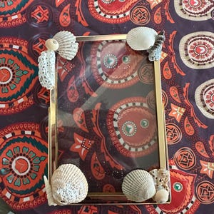 May include: A rectangular gold-coloured picture frame decorated with seashells. The frame is adorned with various types of white seashells and small coral pieces. The background features a patterned fabric with red, orange, and purple paisley designs.