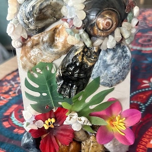 May include: A decorative rectangular object decorated with seashells, small stones, and artificial flowers. The seashells are various colours, including brown, black, and grey. The flowers are red and pink, with green leaves. The object is on a patterned surface.