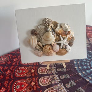 May include: A canvas featuring an arrangement of seashells, including a starfish, in shades of beige, brown, and white. The shells are arranged to create a textured, natural look. The canvas is displayed on a small wooden easel.