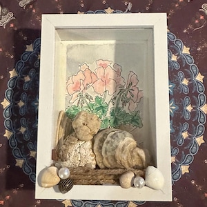 May include: A white framed shadow box with a watercolour painting of pink flowers and green leaves. The box also contains seashells, pearls, and driftwood, creating a coastal theme.