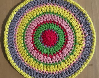 Könnte beinhalten: Ein handgefertigter, kreisförmiger Häkeluntersetzer mit einem lebendigen, konzentrischen Design. Die Mitte ist leuchtend rot, umgeben von Ringen in Grün, Rosa, Gelb, Grau und einem gelben Rand. Der Untersetzer ist wahrscheinlich aus Baumwollgarn.