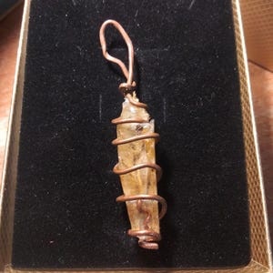 May include: A pendant featuring a light yellow crystal wrapped in copper wire. The crystal is rectangular with a pointed end. The wire is coiled around the crystal and forms a loop at the top for hanging. The pendant is in a black velvet box.