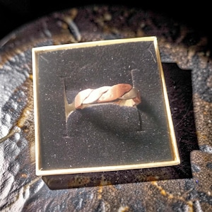 May include: A braided ring with copper and silver tones rests in a small, square box with a black interior. The ring's design features interwoven metal strands, creating a textured appearance. The box is set against a dark, patterned background.