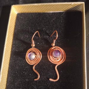 May include: A pair of handcrafted copper wire earrings. Each earring features a spiral design with a small, round, purple gemstone at the center. The earrings have a hook closure and a unique, artistic style.