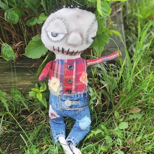 May include: A handmade rag doll with a stitched face, black hair, and a plaid shirt. The doll wears blue denim jeans with patches and white, elongated feet. The doll is posed in a grassy area.