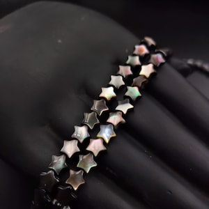 Op de afbeelding: Een zwart armband met stervormige iriserende kralen. De kralen hebben een parelmoer effect, met kleuren als wit, roze en groen. De armband wordt getoond tegen een zwarte achtergrond.