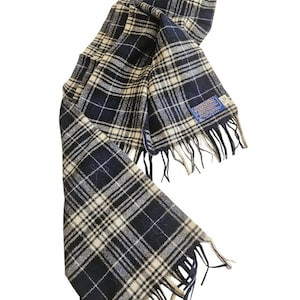 May include: A navy and cream plaid scarf with fringed edges. The scarf is made of a soft material and features a classic plaid pattern. A blue tag is attached to the scarf.