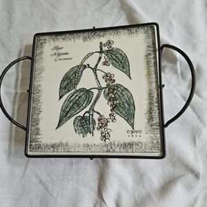 May include: Square ceramic trivet with a black metal frame and handles. The trivet features a botanical illustration of a pepper plant with the text "Piper Nigrum by McCormick" and the year "1955". The design is in shades of green and brown.