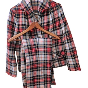 Op de afbeelding: Een vintage geruit pak, bestaande uit een jasje en een bijpassende broek, hangt aan een houten kleerhanger. Het patroon van het pak bevat rode, zwarte, witte en gele lijnen. De jas heeft een reverskraag en zakken. De broek heeft rechte pijpen.