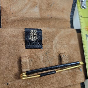 May include: A vintage leather travel case with a gold-toned tie clip. The case is open, revealing a tan suede interior and a black leather exterior. The tie clip is black and gold. The label reads "MARK CROSS ENGLAND".