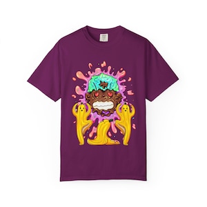 May include: A purple t-shirt featuring a colorful cartoon design. The graphic includes a brown, ice cream-like character with a green and blue head, surrounded by a pink splash. Two yellow banana figures flank a peeled banana.