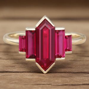 May include: A gold ring with a central, elongated, ruby-coloured gemstone and four smaller, rectangular gems of the same colour. The ring has a geometric design, possibly vintage or Art Deco, set against a wooden background.