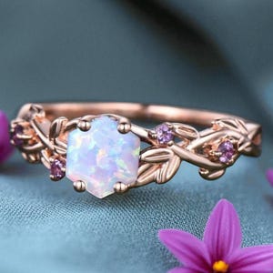 May include: Rose gold ring featuring a hexagonal opal center stone and small purple accent stones. The ring has a floral design with leaves and is set against a teal background. The ring is a piece of jewelry.