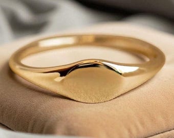 May include: A gold-colored signet ring with a smooth, polished finish. The ring has a rounded band and a flat, oval-shaped face. The ring is displayed on a beige surface, with a soft, blurred background.