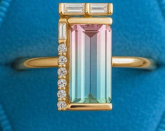 May include: A gold ring with a rectangular, multi-colored gemstone. The gemstone transitions from pink to teal. The ring is accented with small diamonds and baguette-cut stones. The ring is set against a blue velvet background.