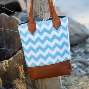 Puede incluir: Un bolso tote de color azul y blanco con estampado de chevron, con asas y ribetes de cuero marrón. El bolso está colgado de una valla de cadena.