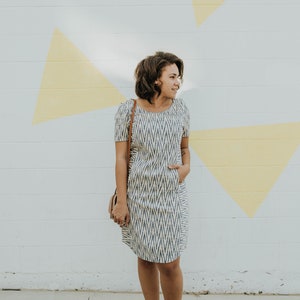 Könnte beinhalten: Eine Frau in einem weiß-blau gemusterten Kleid mit einer braunen Tasche und gelben Schuhen steht vor einer weißen Wand mit gelben geometrischen Formen.