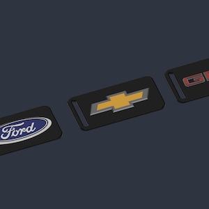 May include: Three rectangular black keychains featuring automotive logos. The first displays the blue and white Ford oval logo, the second shows the gold and silver Chevrolet bowtie emblem, and the third features the red and silver GMC lettering.