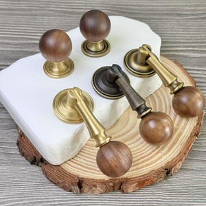 Può includere: Una collezione di ferramenta per mobili in legno e ottone, tra cui pomelli e maniglie. I pomelli hanno una parte superiore rotonda in legno e una base in ottone. Le maniglie presentano una sfera di legno all'estremità di una maniglia in ottone o metallo scuro.