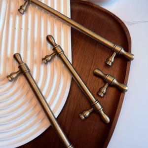 以下が含まれることがあります： T字型のデザインのアンティーク真鍮キャビネットプルをいくつかクローズアップ。プルは、白いリブ付きの表面と濃い木製のトレイに配置されています。ハードウェアはヴィンテージの美学を持っています。