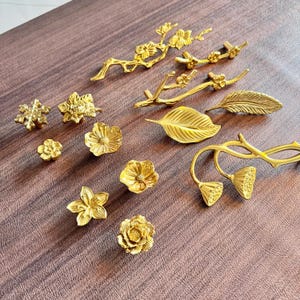 May include: A selection of gold-coloured decorative hardware, including drawer pulls and knobs. The hardware features floral and botanical designs, such as flowers, leaves, and branches. The pieces are arranged on a brown surface.