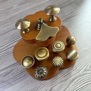 May include: A collection of antique brass cabinet knobs and pulls in various designs, including round, floral, and geometric shapes. The hardware is displayed on a wooden tiered stand, showcasing the different styles and finishes.