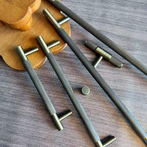 May include: A collection of dark gray cabinet pulls and a small knob. The pulls have a textured, diamond-patterned grip and a T-shaped handle. The hardware is arranged on a wooden surface and a gray fabric background.