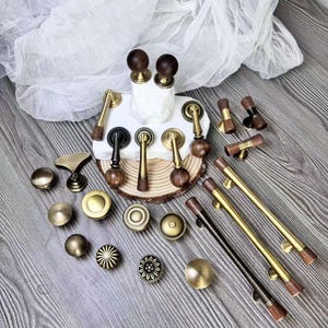 Może przedstawiać: Kolekcja różnych okuć do szafek, w tym gałek i uchwytów, w wykończeniach mosiężnych, czarnych i drewnianych. Okucia mają okrągłe, kwiatowe i prętowe wzory. Niektóre elementy mają ciemnobrązowe wykończenie z usłojeniem drewna.