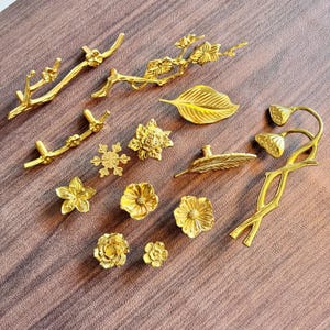 Może przedstawiać: Kolekcja złotych elementów dekoracyjnych, w tym uchwytów i gałek do szuflad. Asortyment obejmuje wzory kwiatowe, liściaste i gałęzi. Okucia ułożone są na brązowej powierzchni, prezentując różne kształty i rozmiary.