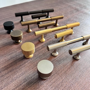 Op de afbeelding: Een verzameling kastbeslag, waaronder diverse knoppen en stanggrepen. De knoppen zijn rond met een geribbeld ontwerp en de grepen zijn T-vormig met een geribbelde stang. Afwerkingen zijn zwart, goud en zilver. Voor meubels.