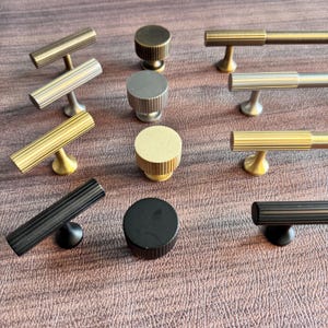 Może przedstawiać: Kolekcja różnych okuć do szafek, w tym gałek i uchwytów. Okucia są dostępne w kolorach złotym, srebrnym i czarnym. Gałki są okrągłe, a uchwyty mają kształt pręta z żebrowanymi detalami.