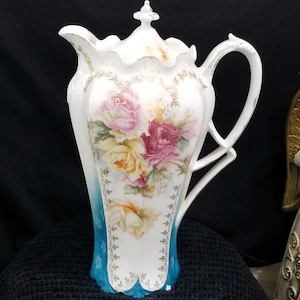 May include: A white porcelain pitcher with a lid, featuring a floral design of pink, yellow, and red roses. The pitcher has a blue base and a decorative handle. Gold trim accents the edges and floral details.